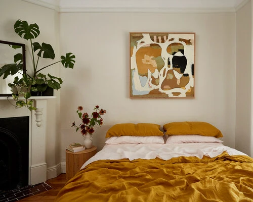 Bedroom interior with a mustard-colored bed, abstract art, and potted plants creating a cozy and inviting home atmosphere.