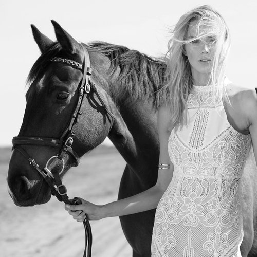 Monochrome portrait of a woman with long blonde hair standing next to a horse in an outdoor environment with a sandy surface.