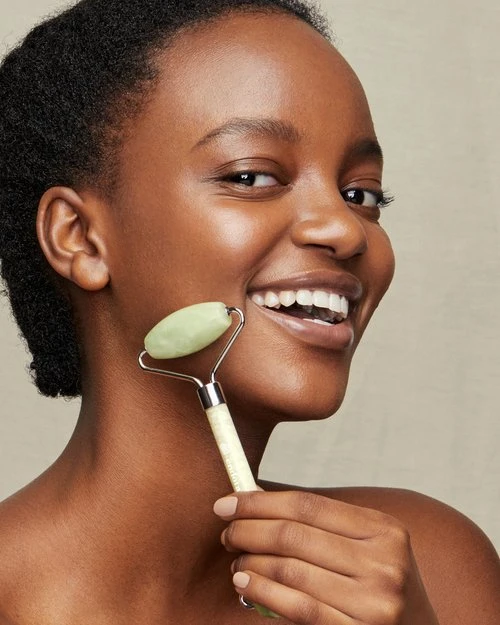 Smiling African American woman uses a jade facial roller on her face in an eye-level shot. Beauty and skincare concept.