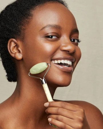Smiling African American woman uses a jade facial roller on her face in an eye-level shot. Beauty and skincare concept.