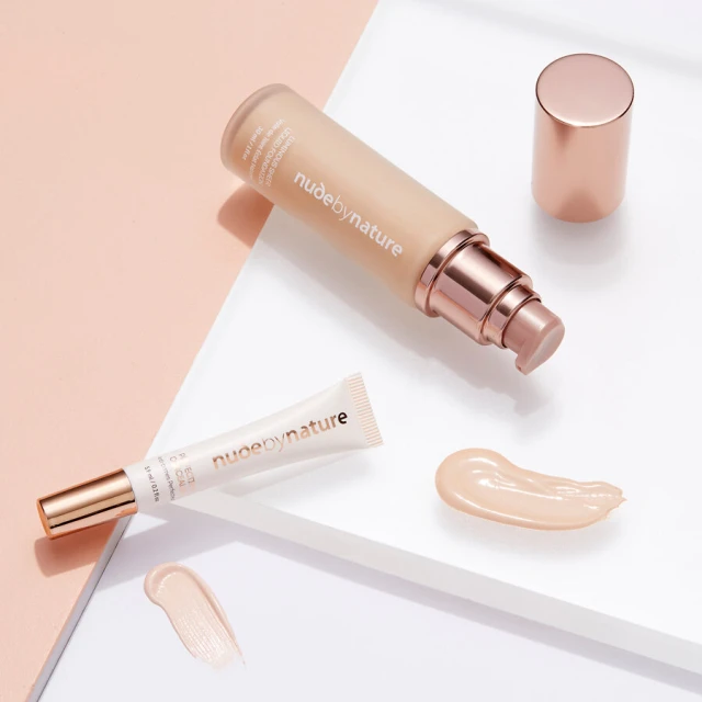 Nude by Nature foundation bottle with lid, makeup tube, and swatches on a white surface against a light pink and white background.