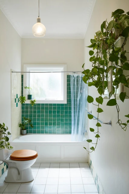 Bathroom interior featuring a white bathtub, green tiles, toilet, trailing plants, and a window providing natural light.
