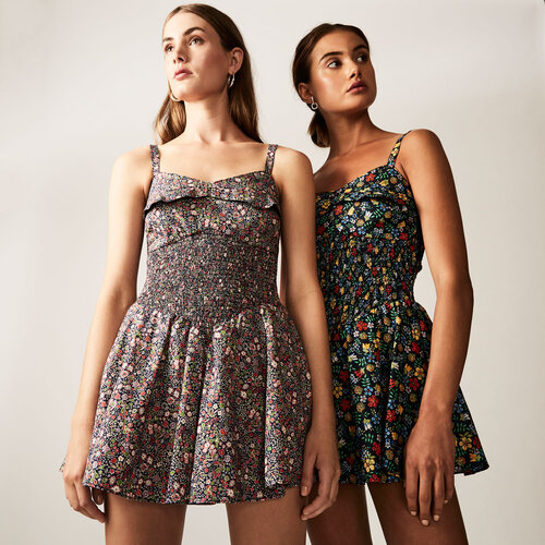Two women in floral mini dresses pose in a studio. The dresses have dark backgrounds with a colorful scattered flower pattern.