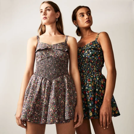 Two women in floral mini dresses pose in a studio. The dresses have dark backgrounds with a colorful scattered flower pattern.