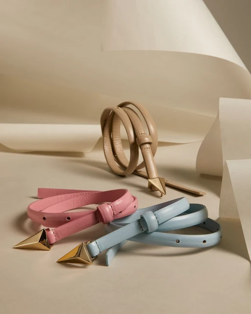Stylish belts in beige, pink, and light blue colors with gold hardware accents arranged artfully on a cream-colored surface and backdrop.