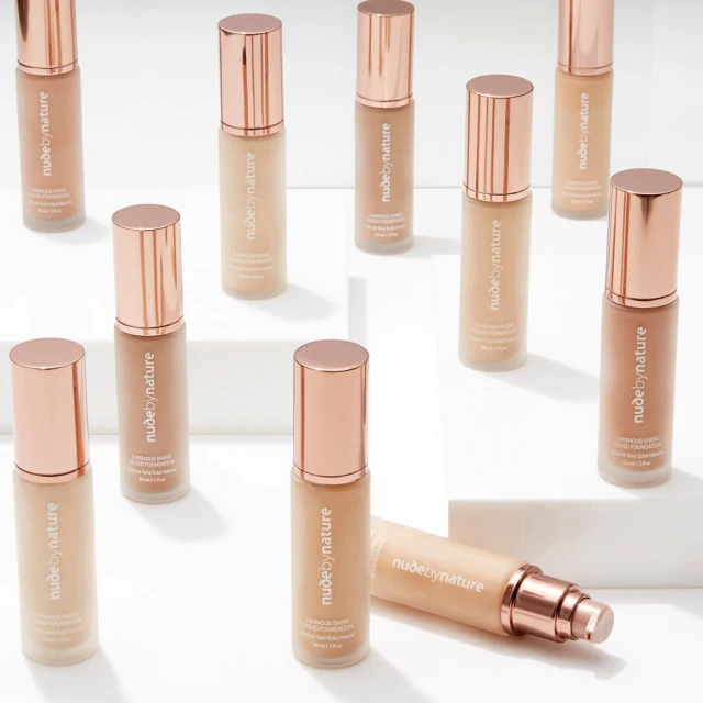 Multiple bottles of Nude by Nature foundation are arranged on a white surface, displaying the brand's makeup product in an organized manner.