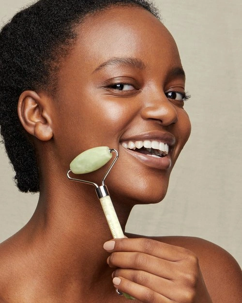 Close-up portrait of a Black woman using a green jade facial roller on her cheek against a neutral background.