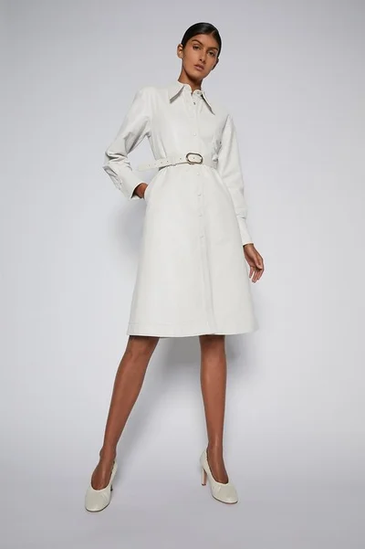 Model in a white belted dress and matching heels standing against a neutral studio background