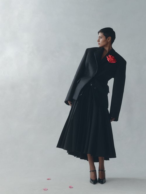 Elegant woman in a black suit and full skirt poses in a bright photo studio with scattered red flower petals.