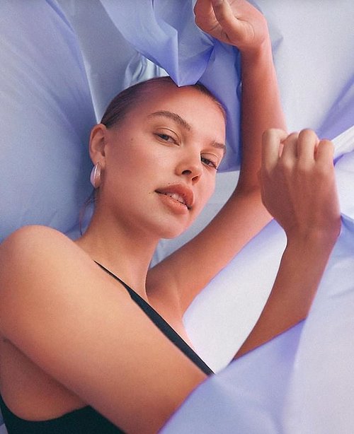 Portrait of a fair-skinned adult woman with slicked-back hair, wearing a black tank top, posing with periwinkle fabric around her.