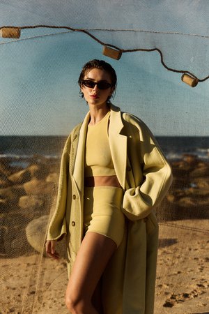 Woman in yellow shorts and top with a tan trench coat, sunglasses, short hair poses in front of a sandy beach with ocean background.