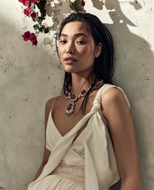 Asian woman in a draped white dress and ruby necklace poses in front of a textured wall with floral accents, showcasing elegance.