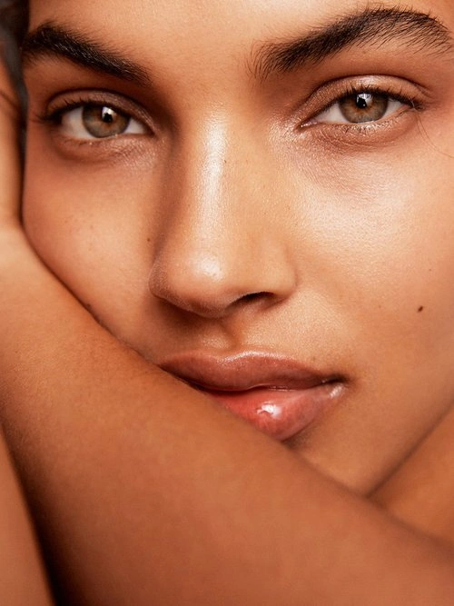 Close-up portrait of a woman resting her face against her arm, highlighting natural skin texture and hazel eyes.