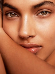 Close-up portrait of a woman resting her face against her arm, highlighting natural skin texture and hazel eyes.