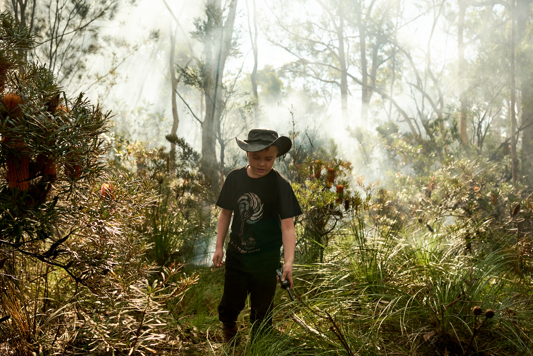 Young boy in a hat walking through a forest with tall grasses, plants, and trees with smoke in the background in a natural setting.