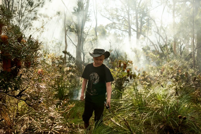 A young boy in a black t-shirt and bush hat walks through a smoky bushland forest in Sydney captured by Alex Brunton.