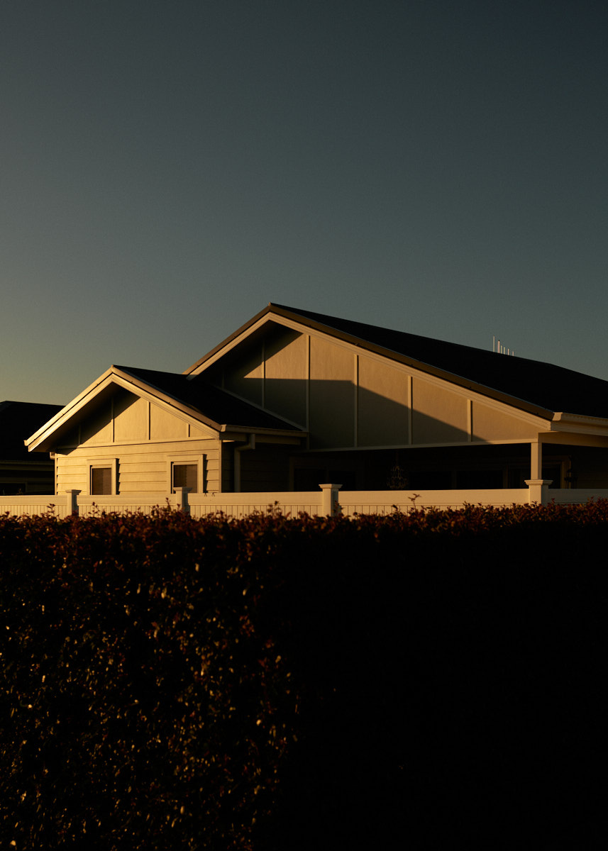 Exterior view of a modern home at sunset featuring clean architectural lines, warm lighting, and a dark hedge in the foreground.