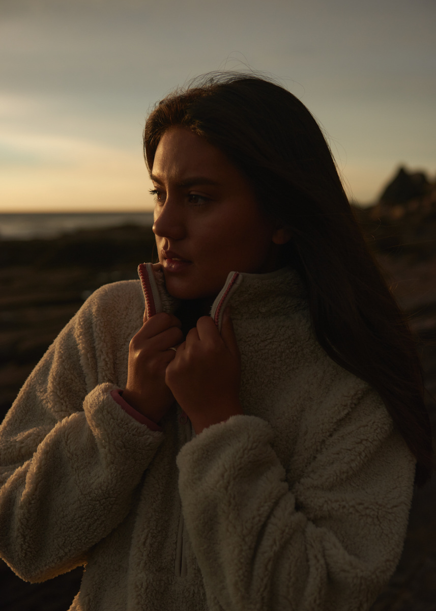 A young woman in a fleece jacket stands outdoors near a beach with a serious expression, gazing toward the horizon at sunset.