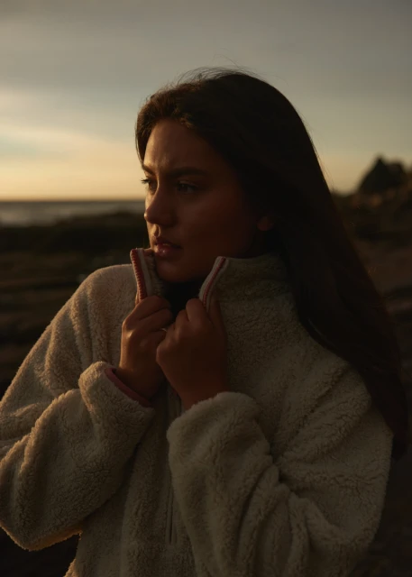 A young woman in a fleece jacket stands outdoors near a beach with a serious expression, gazing toward the horizon at sunset.