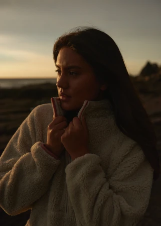 A young woman in a fleece jacket stands outdoors near a beach with a serious expression, gazing toward the horizon at sunset.