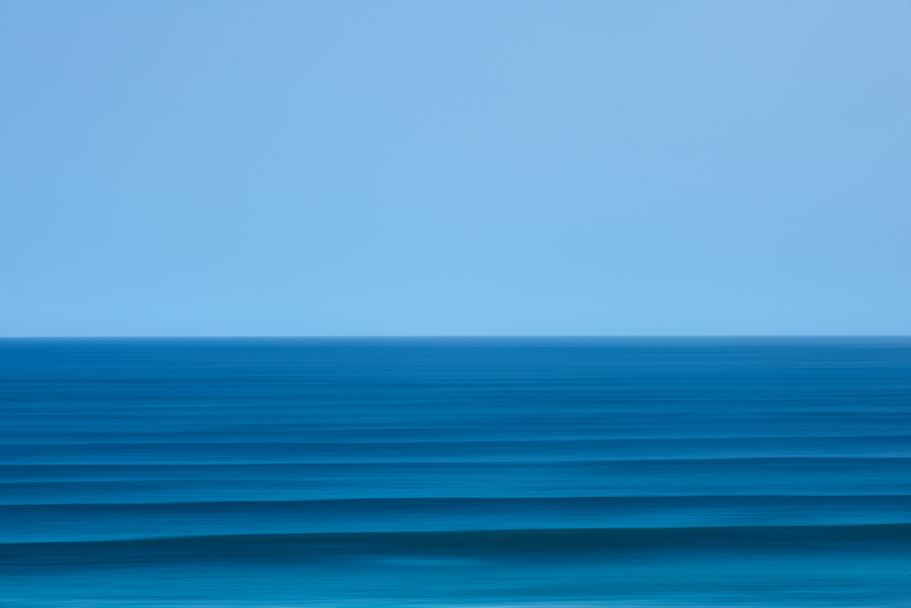 Ocean horizon with clear blue sky and teal waves creating a minimalist, serene seascape in a horizontal, panoramic composition.