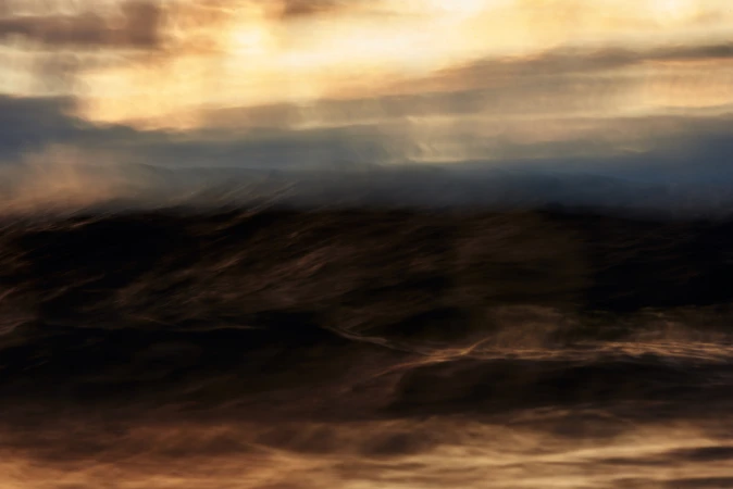 Abstract landscape with dark, blurred land under a cloudy, golden sky. Long exposure creates a dreamy, atmospheric vista.