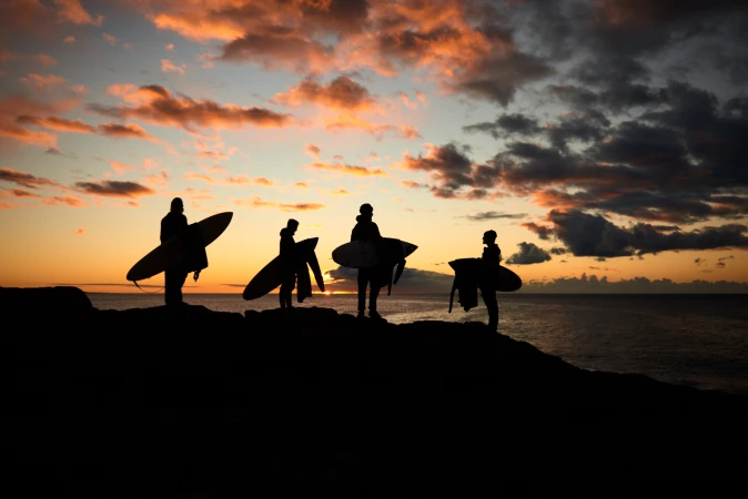 Silhouetted surfers standing on a dark beach with surfboards at sunset, ocean and cloudy sky in background, wide angle.