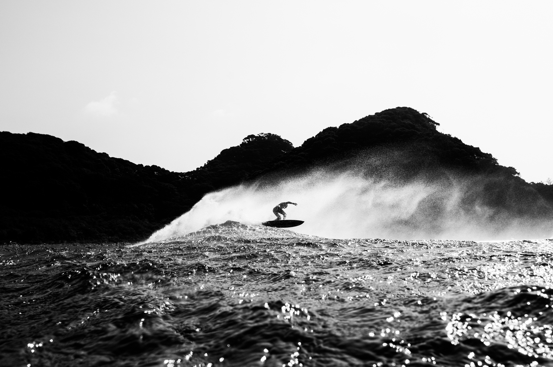 Black and white shot of a surfer riding a wave with forested hills in the background under a bright sky, captured in an eye-level shot.
