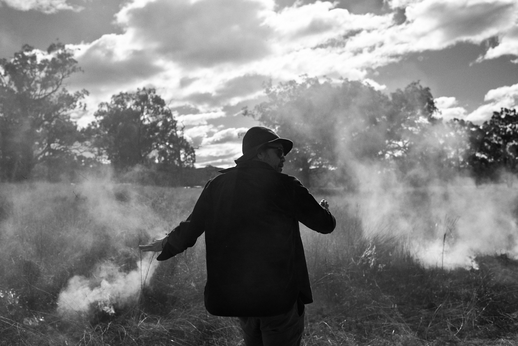 Black and white outdoor portrait of an adult wearing a hat and jacket, standing in a field surrounded by haze and trees in the background.