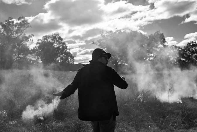 Black and white outdoor portrait of an adult wearing a hat and jacket, standing in a field surrounded by haze and trees in the background.