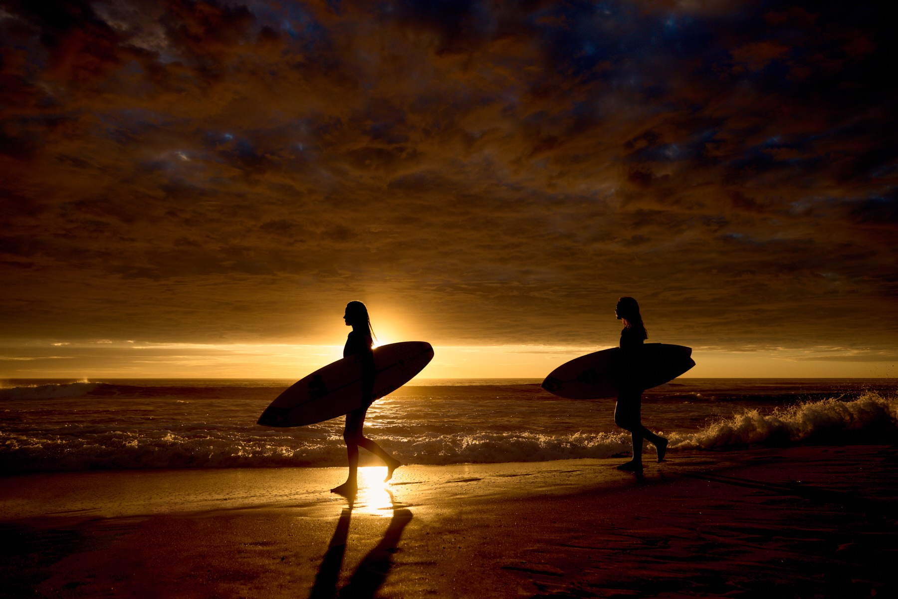 Two silhouetted women carrying surfboards walk on a beach at sunset with orange and dark clouds in the background.