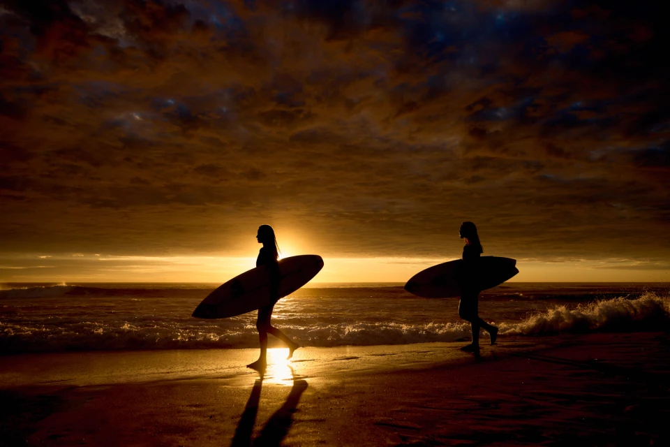 Two silhouetted women carrying surfboards walk on a beach at sunset with orange and dark clouds in the background.