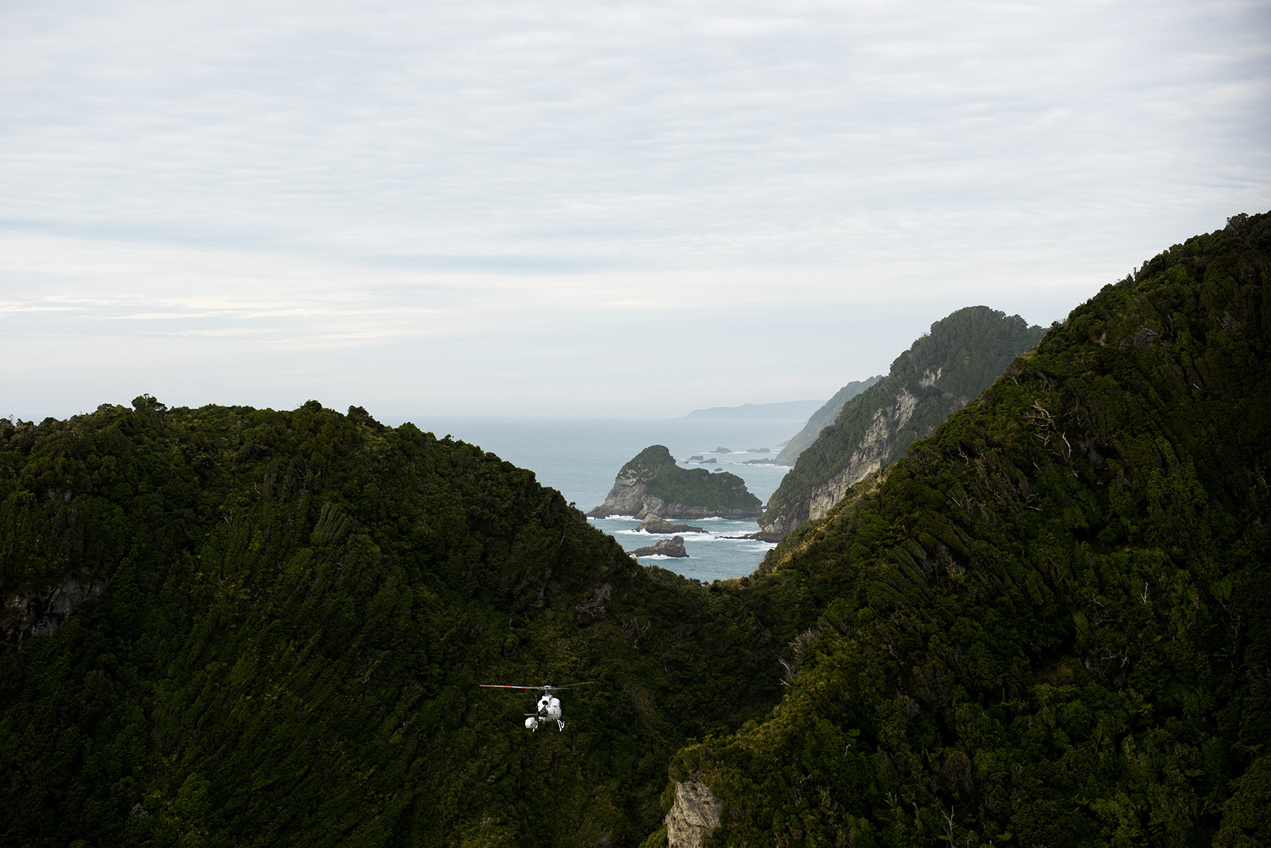 Aerial view of a helicopter flying through a mountain pass with a forested landscape and ocean in the background, under an overcast sky.