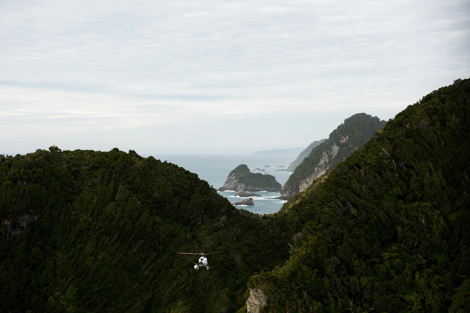 Aerial view of a helicopter flying through a mountain pass with a forested landscape and ocean in the background, under an overcast sky.