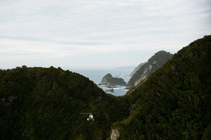 Aerial view of a helicopter flying through a mountain pass with a forested landscape and ocean in the background, under an overcast sky.