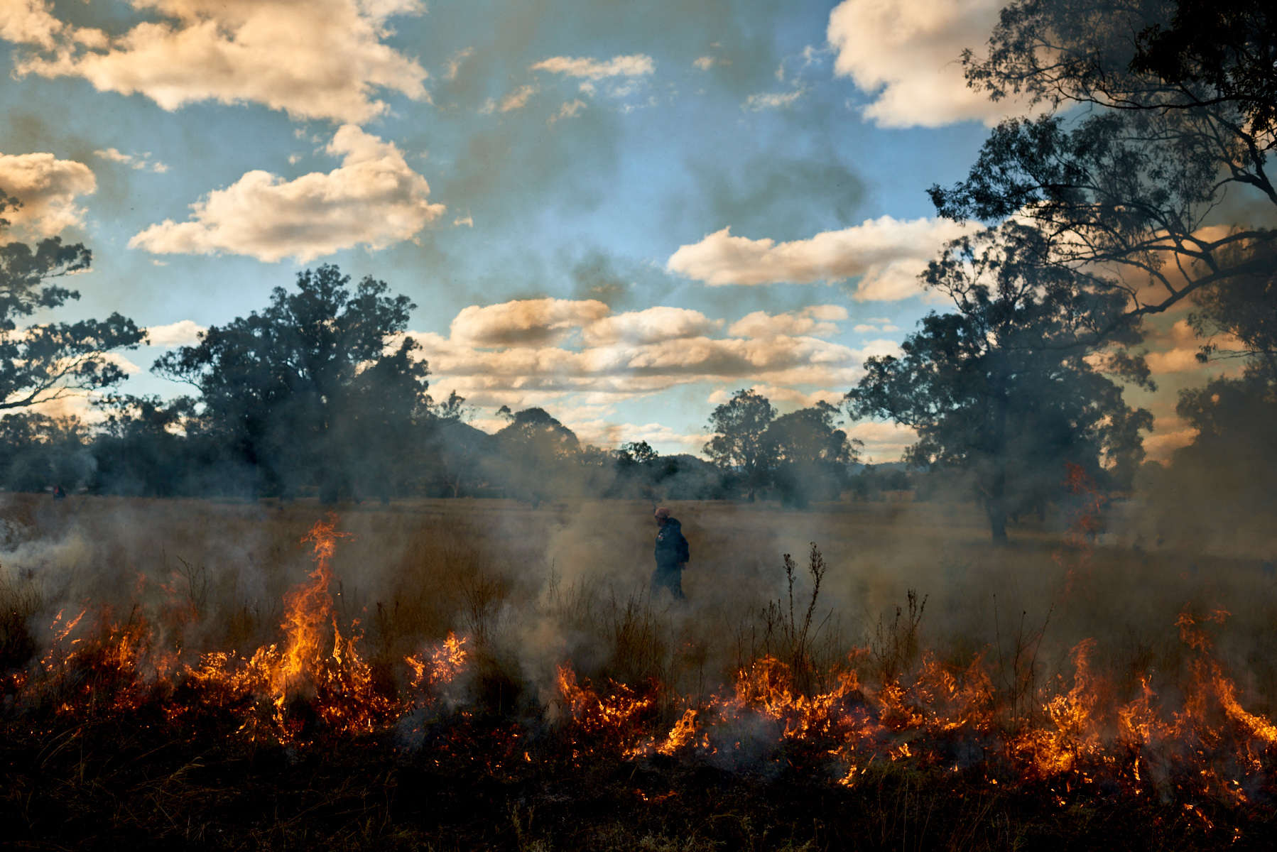 Controlled burn in a grassy field with a single adult standing in the distance amid the smoke and flames, with scattered trees and clouds.
