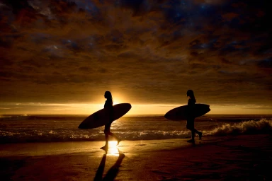 Two surfers carrying surfboards along a sandy beach at sunset in silhouette against a dramatic dark cloud sky