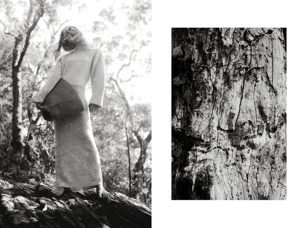 Monochrome fashion shot of a bare-footed model in a long dress holding a square bag, posed in a forest against a tree trunk background.