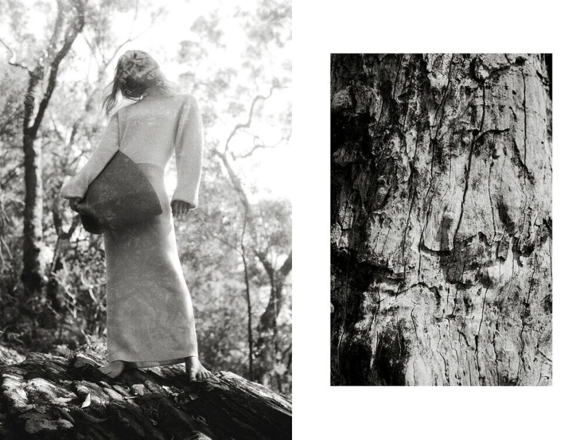 Monochrome fashion shot of a bare-footed model in a long dress holding a square bag, posed in a forest against a tree trunk background.