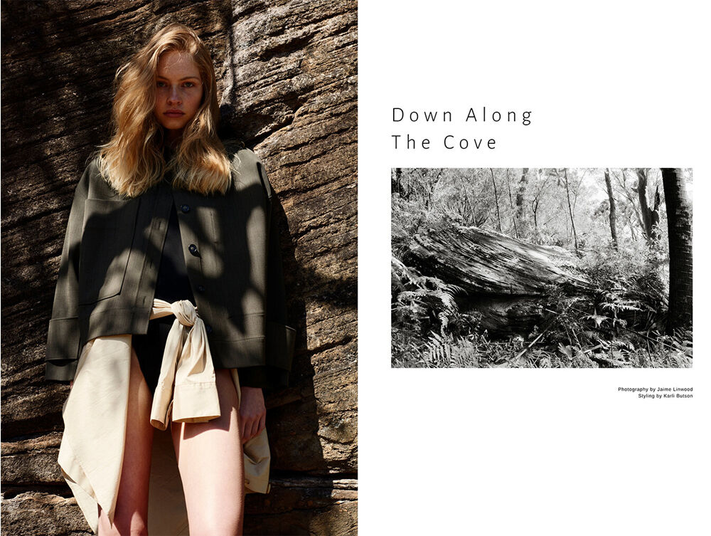 A young woman poses in a shaded outdoor setting, wearing a dark top, dark jacket, and light-colored skirt, near a textured rock formation.