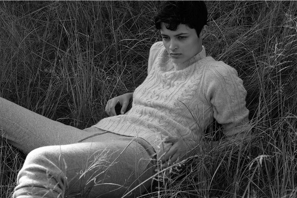Woman in a knit sweater and pants sitting in a field of tall grass in black and white, looking away with a thoughtful expression.
