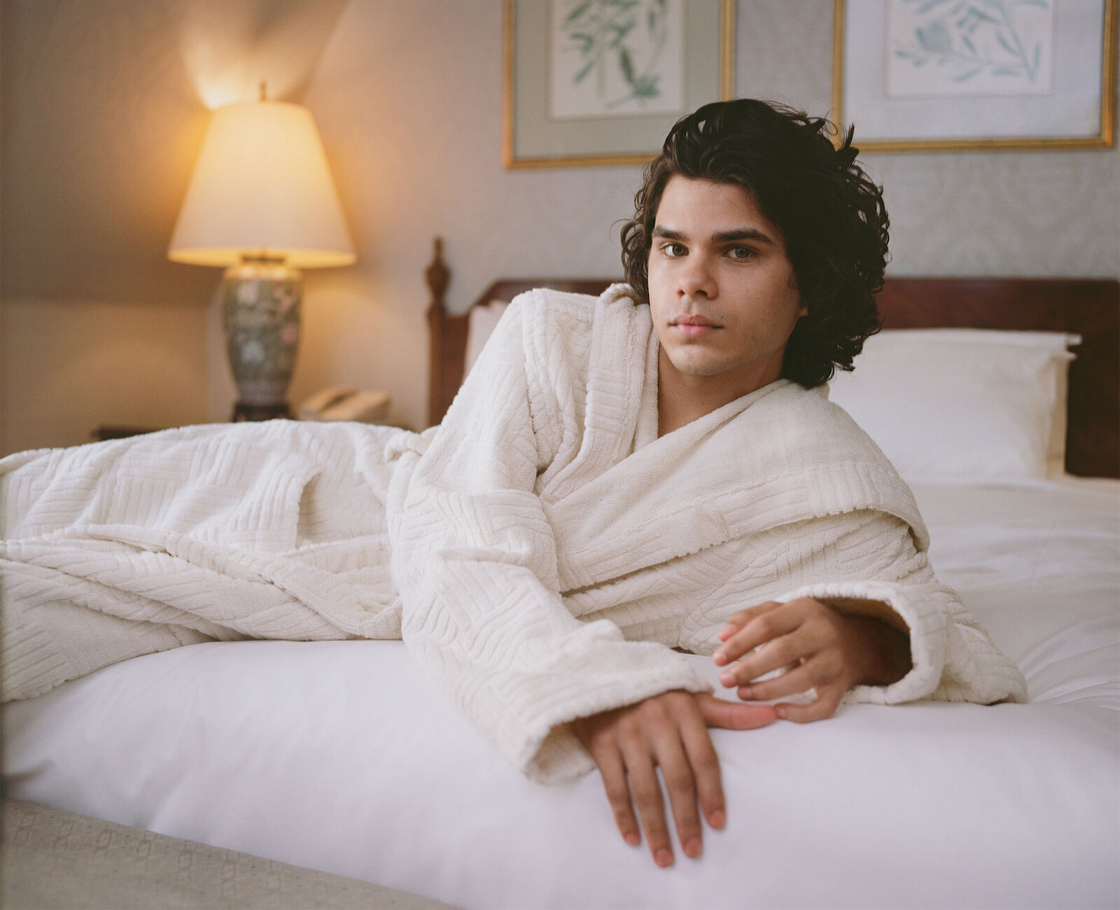Man in a white bathrobe lying on a white bed, illuminated by a nearby lamp, with paintings hanging in the background in a bedroom setting.