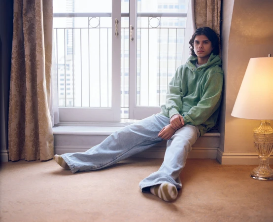 Young man in a green hoodie and light blue jeans sitting on a windowsill by a window with sheer curtains.