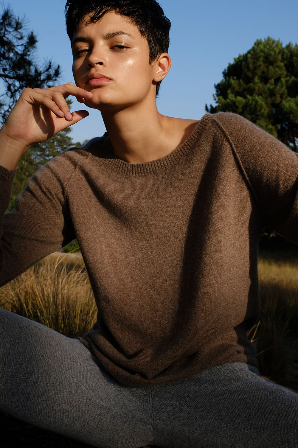 Adult model with short dark hair poses outdoors in a tan sweater and grey leggings, illuminated by natural sunlight in a tranquil setting.