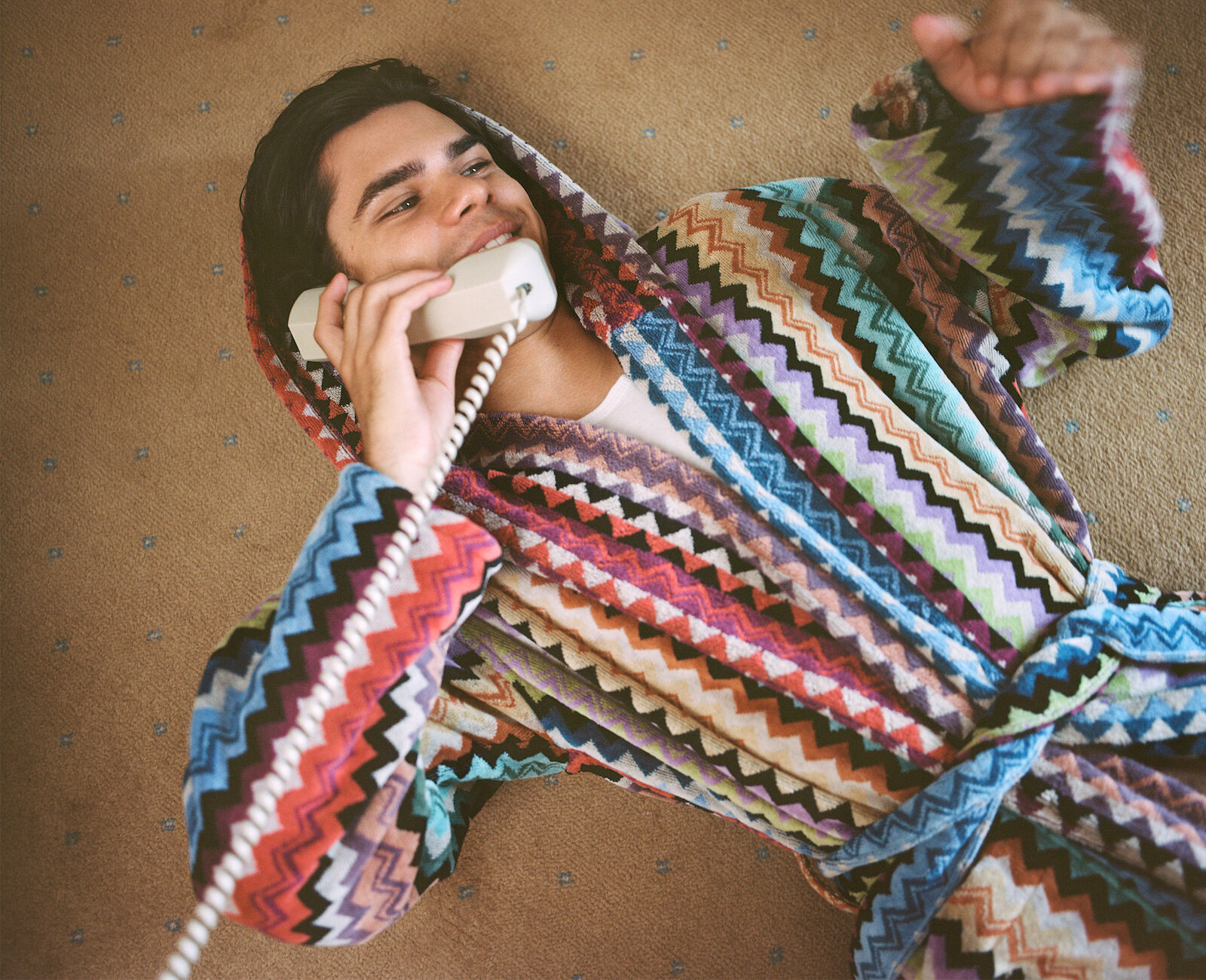 Young man in a colorful chevron-patterned robe lies on a carpet, holding a vintage telephone, smiling. Indoor scene, top-down shot.