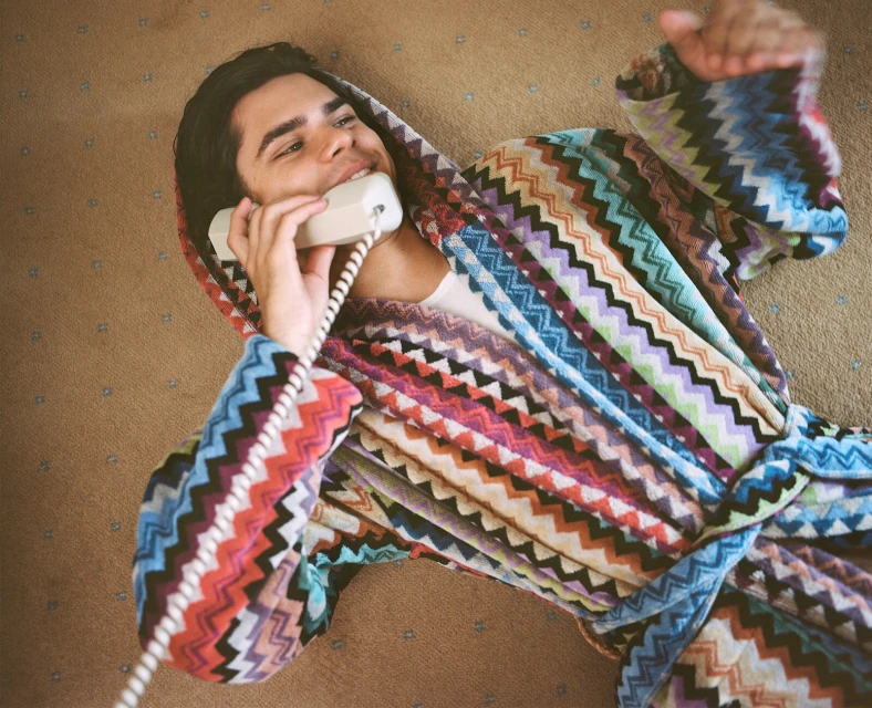 Young man in a colorful chevron-patterned robe lies on a carpet, holding a vintage telephone, smiling. Indoor scene, top-down shot.