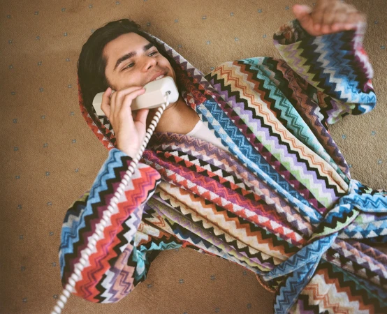 Young man in a colorful chevron-patterned robe lies on a carpet, holding a vintage telephone, smiling. Indoor scene, top-down shot.