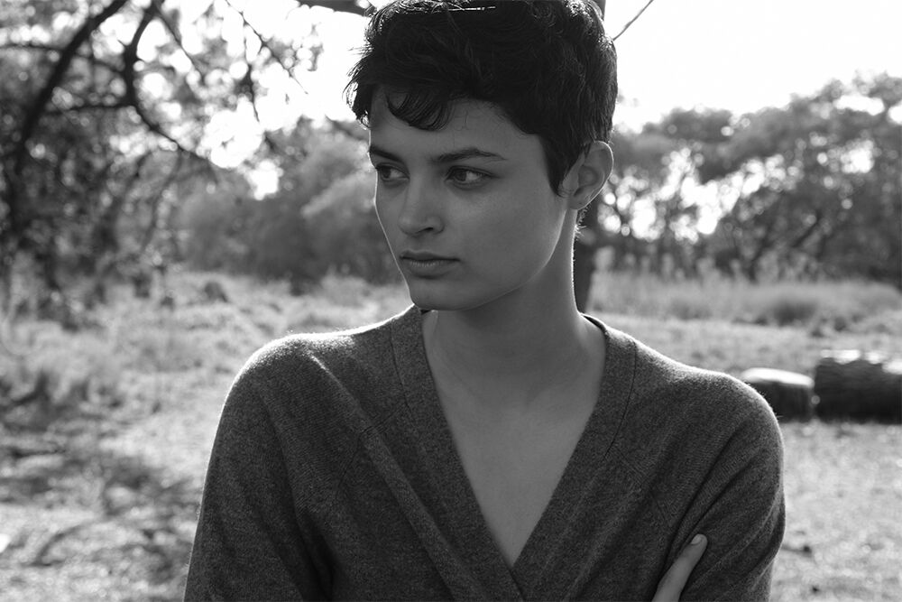 Black and white portrait of an adult female with short, dark hair wearing a grey sweater standing outside with trees in the background.