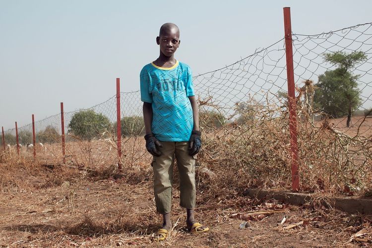 Boy standing in rural setting near fence line, wearing a blue patterned t-shirt, brown pants and yellow sandals.