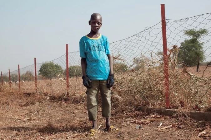 Boy standing in rural setting near fence line, wearing a blue patterned t-shirt, brown pants and yellow sandals.